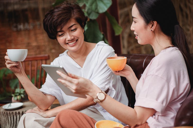 Positive Asian Friends Browsing Tablet And Drinking Coffee