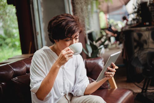 Free Focused Asian female in trendy clothes drinking cup of beverage and looking at screen of tablet Stock Photo