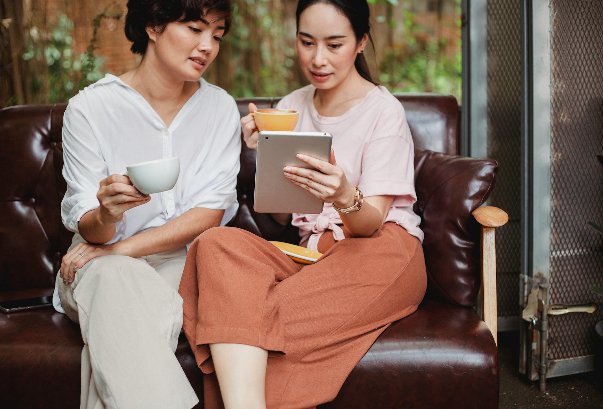 Calm Asian friends sitting with cup of beverage and looking at screen of tablet