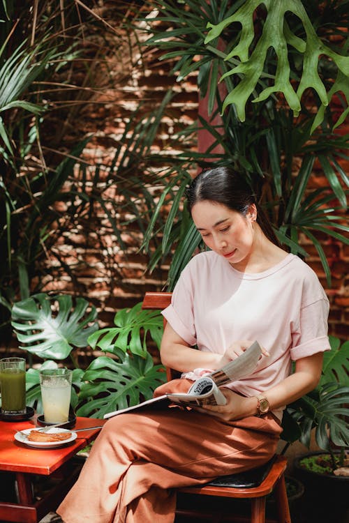 Calm Asian woman in casual clothes sitting in cafe and reading interesting magazine against green plants on brick wall