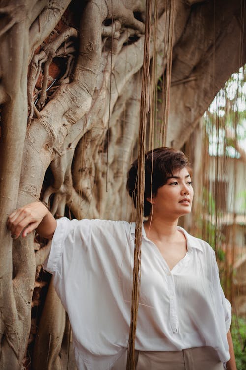 Pensive Asian female with hairstyle in stylish white blouse leaning on aged tree in daytime