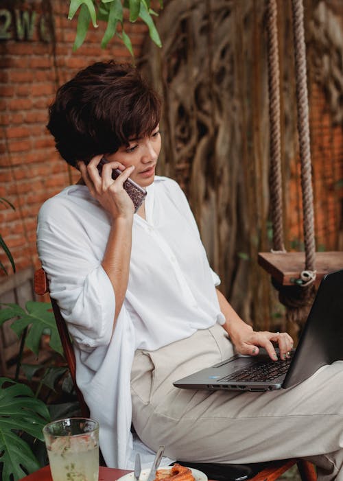 Nachdenkliche Asiatische Frau, Die Laptop Verwendet Und Auf Smartphone Spricht