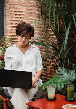 East Asian woman in casual attire working on a laptop in a relaxed café environment, surrounded by greenery.