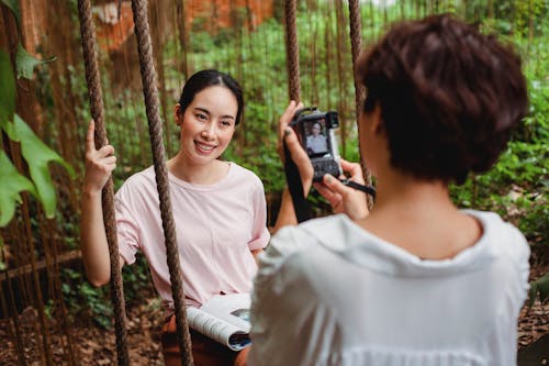 Back view of unrecognizable female taking picture of smiling Asian female sitting on swing in green garden in daytime