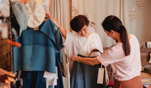 Unrecognizable woman showing clothes to cheerful Asian girlfriend in shop