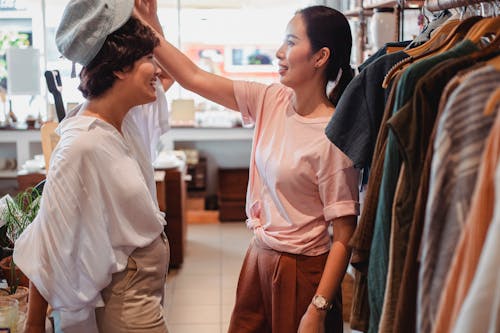 Young smiling ethnic female buyer putting head wear on positive partner while spending time in clothing store
