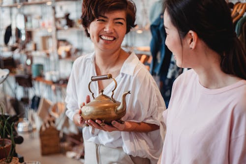 Crop cheerful ethnic female shoppers with oriental kettle speaking in local shop while spending time together and looking at each other