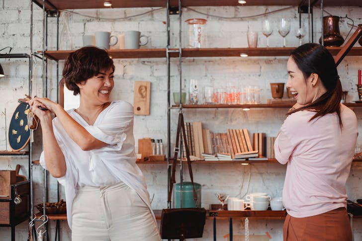 Young ethnic female buyers interacting while laughing near shelves with kitchen items and looking at each other in store