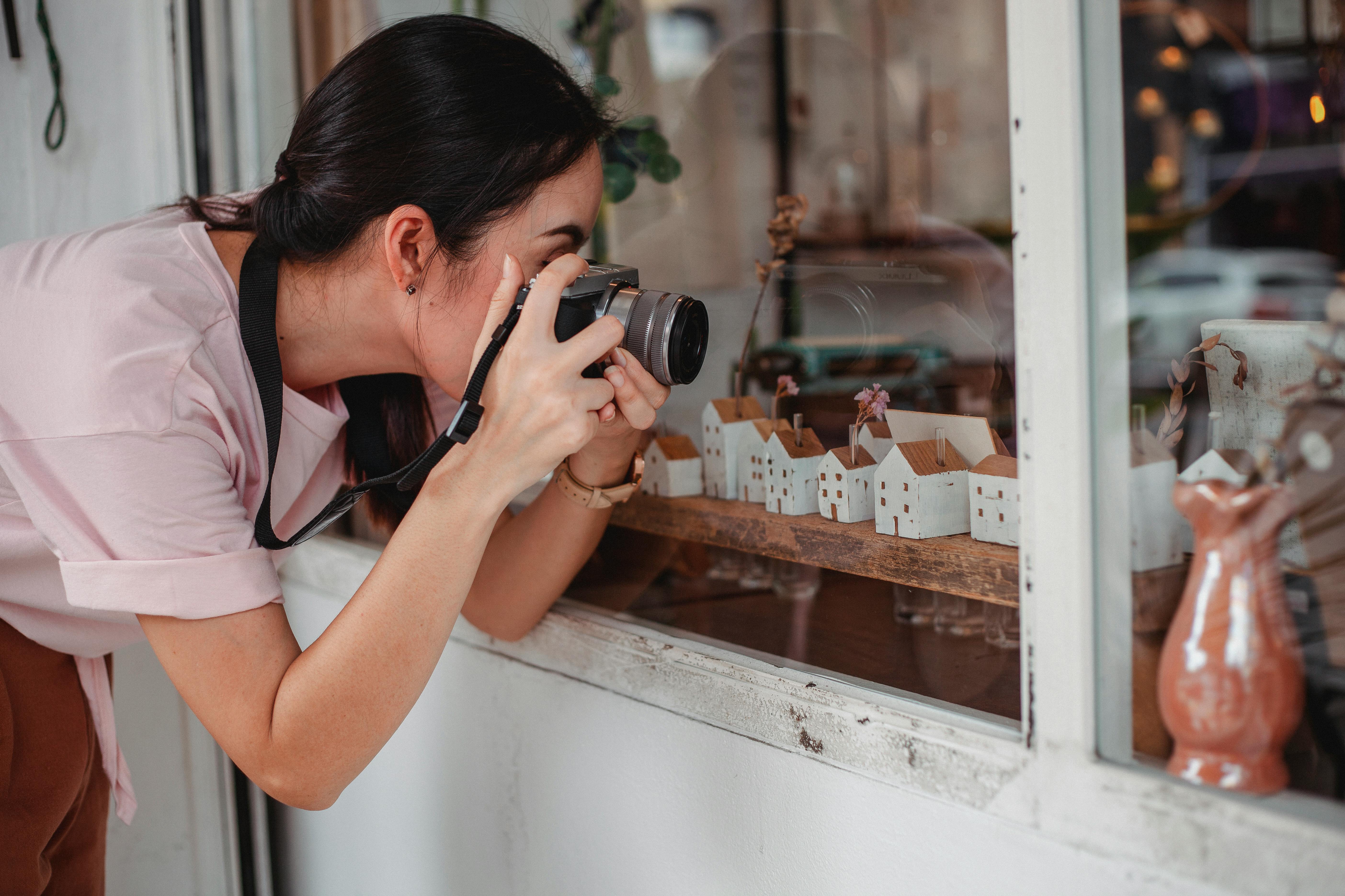 Anonymous woman taking photo of decorative houses on camera near glass wall of urban shop