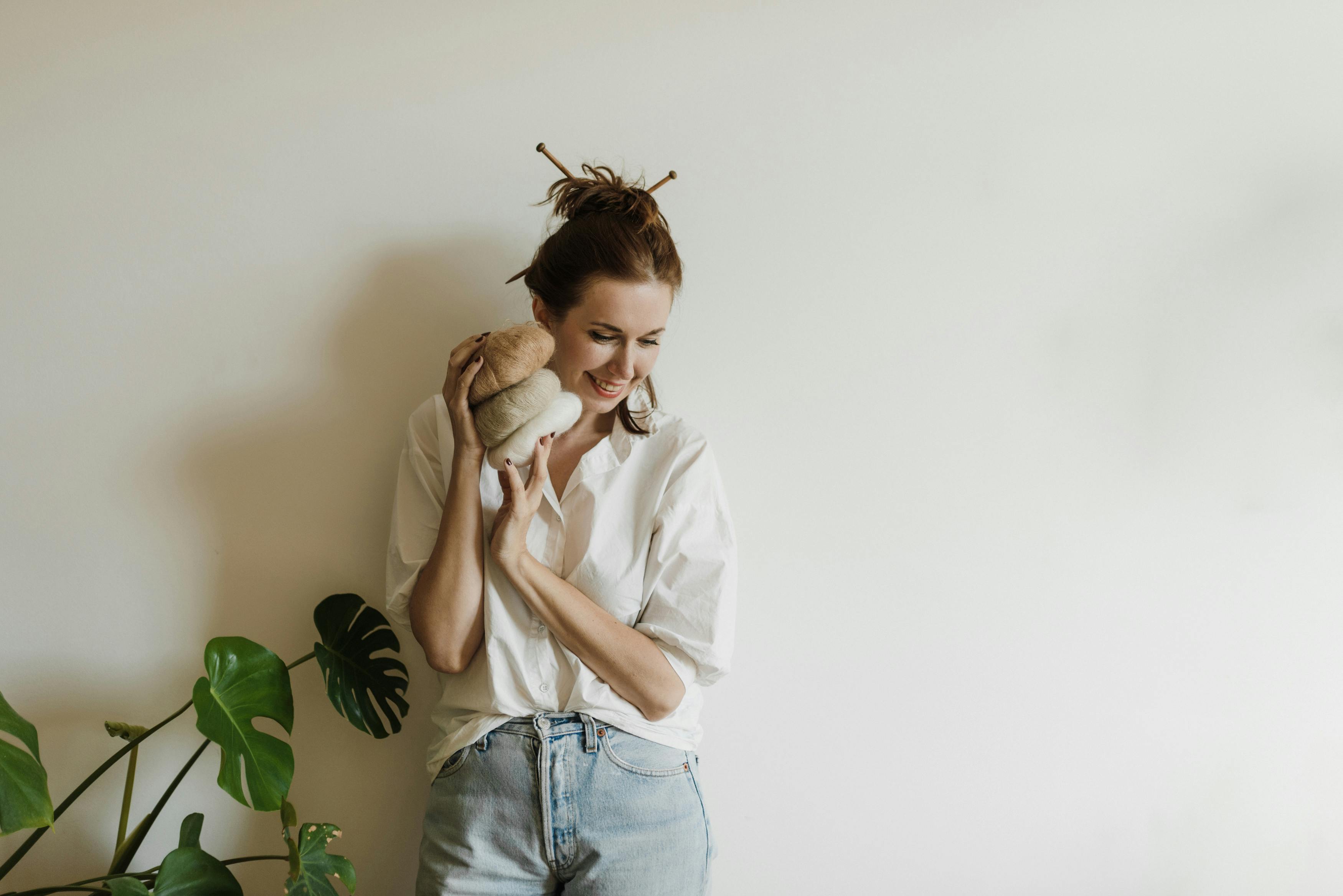 Young woman smiling indoors holding yarn rolls against a white background.