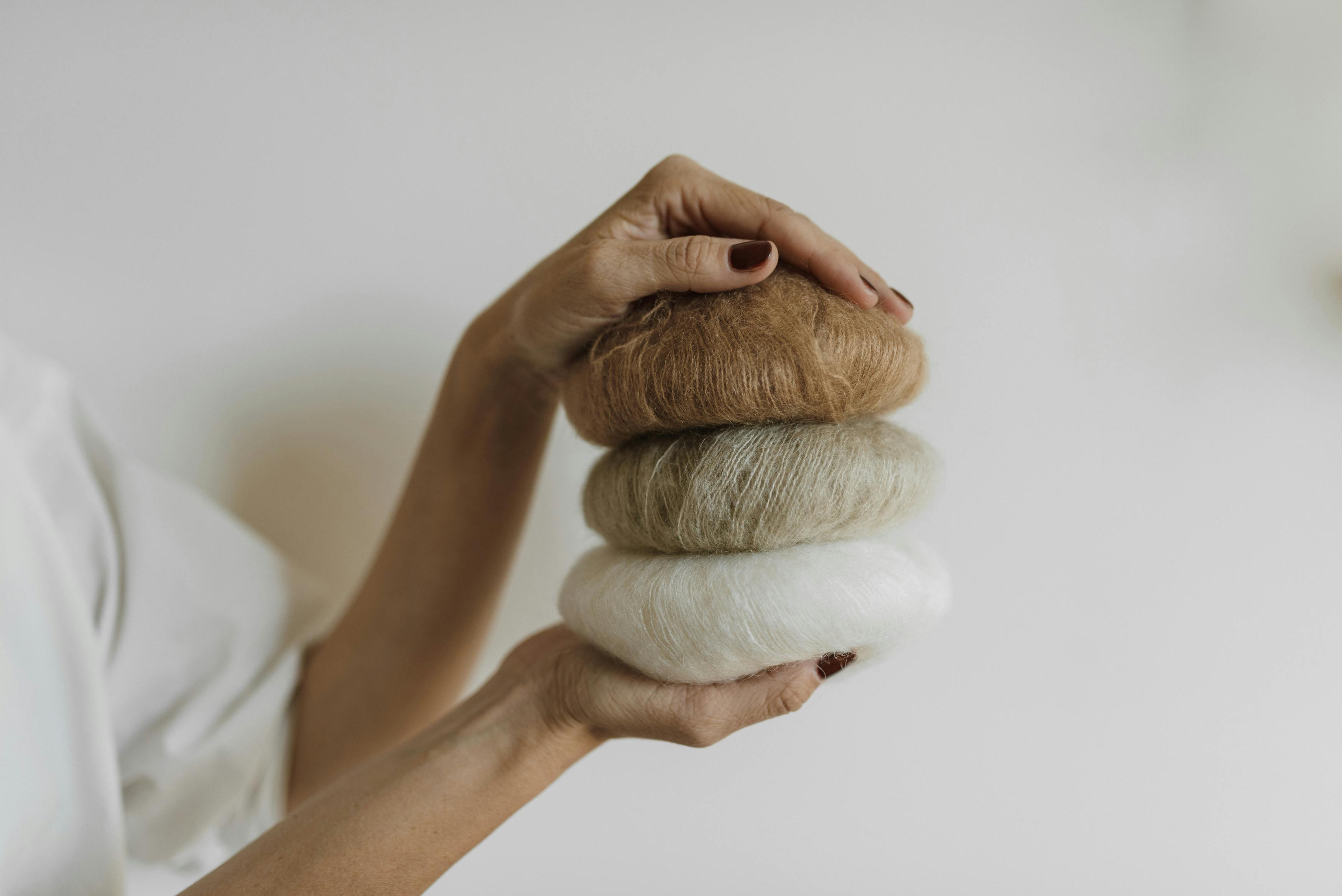A close-up of hands holding stacked colorful yarn balls in a neutral studio setting.