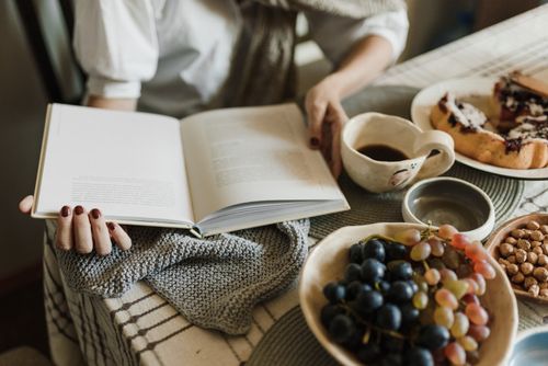 Free Woman reading a book while enjoying coffee, grapes, and pastries in a cozy setting. Stock Photo