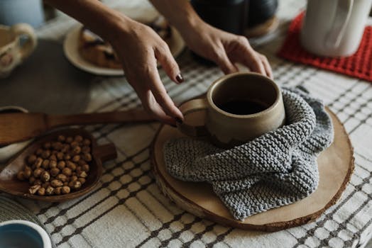 Hands arranging a coffee mug on a knitted cloth with nuts on a wooden board.
