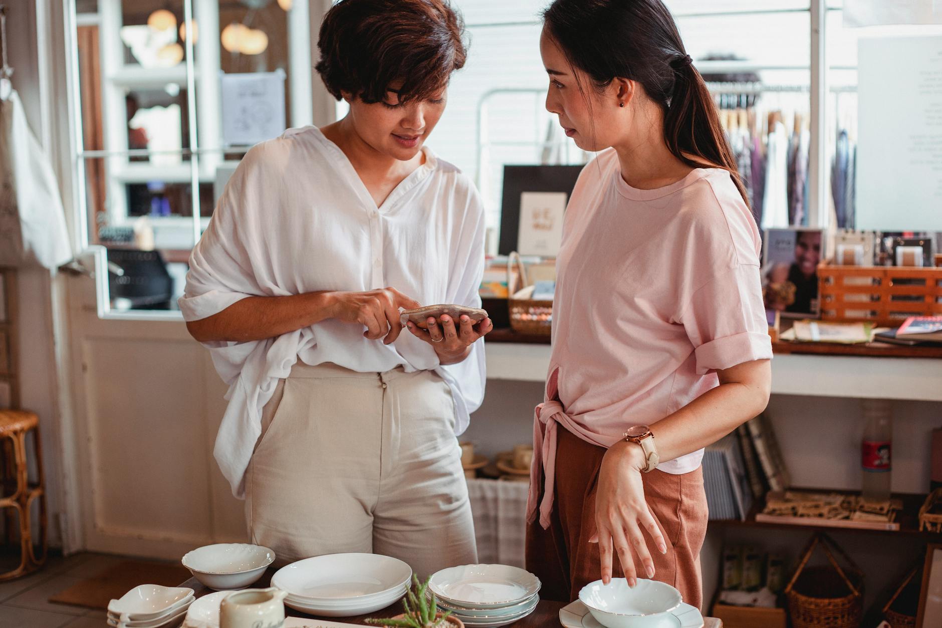 Two women browse ceramics in a boutique using a smartphone, shopping for elegant dishware.