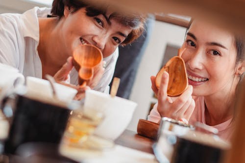 Crop happy Asian female friends smiling while demonstrating wooden kitchenware during shopping in store