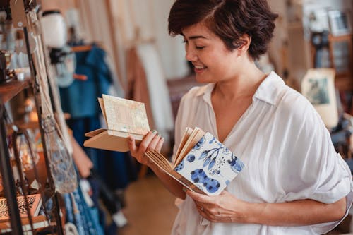 Free Side view of crop young happy Asian lady choosing various notepads while standing near shelf with stylish goods in vintage boutique Stock Photo