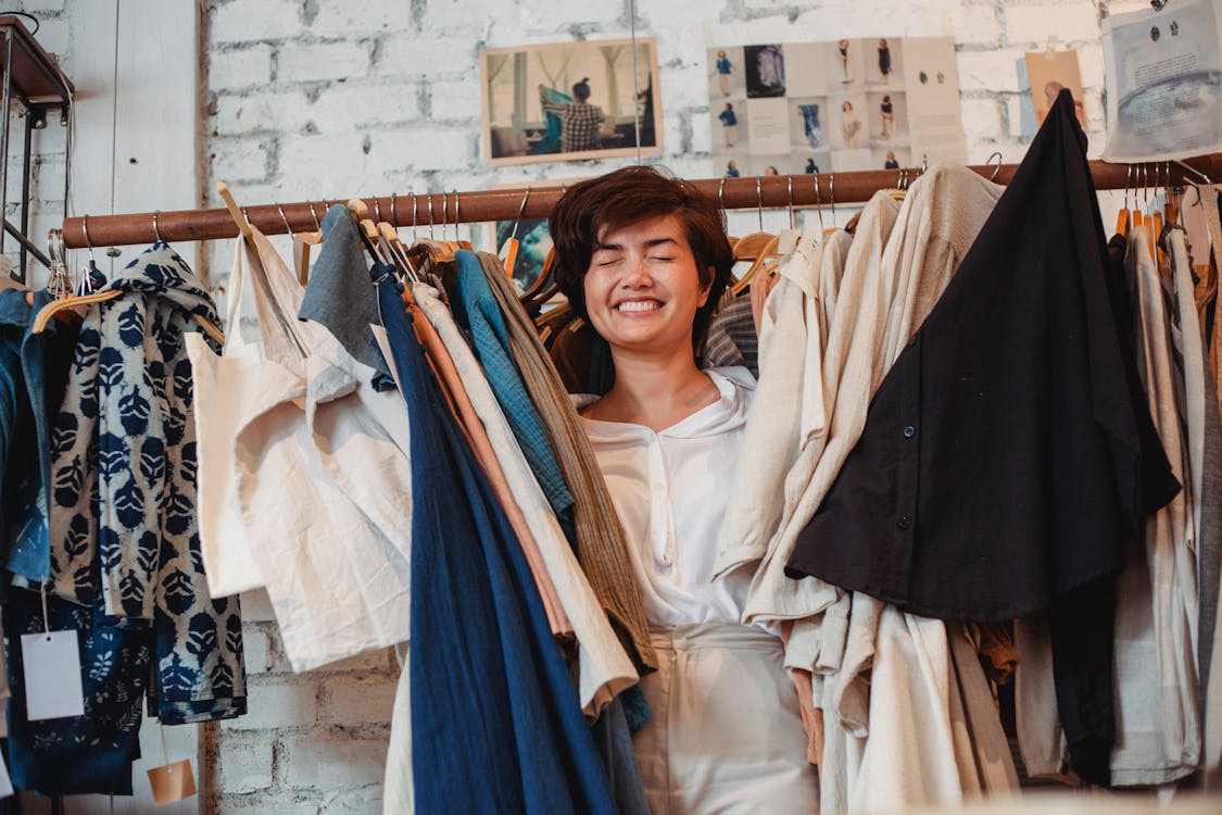 Cheerful Asian female customer standing among hanging clothes in store and smiling