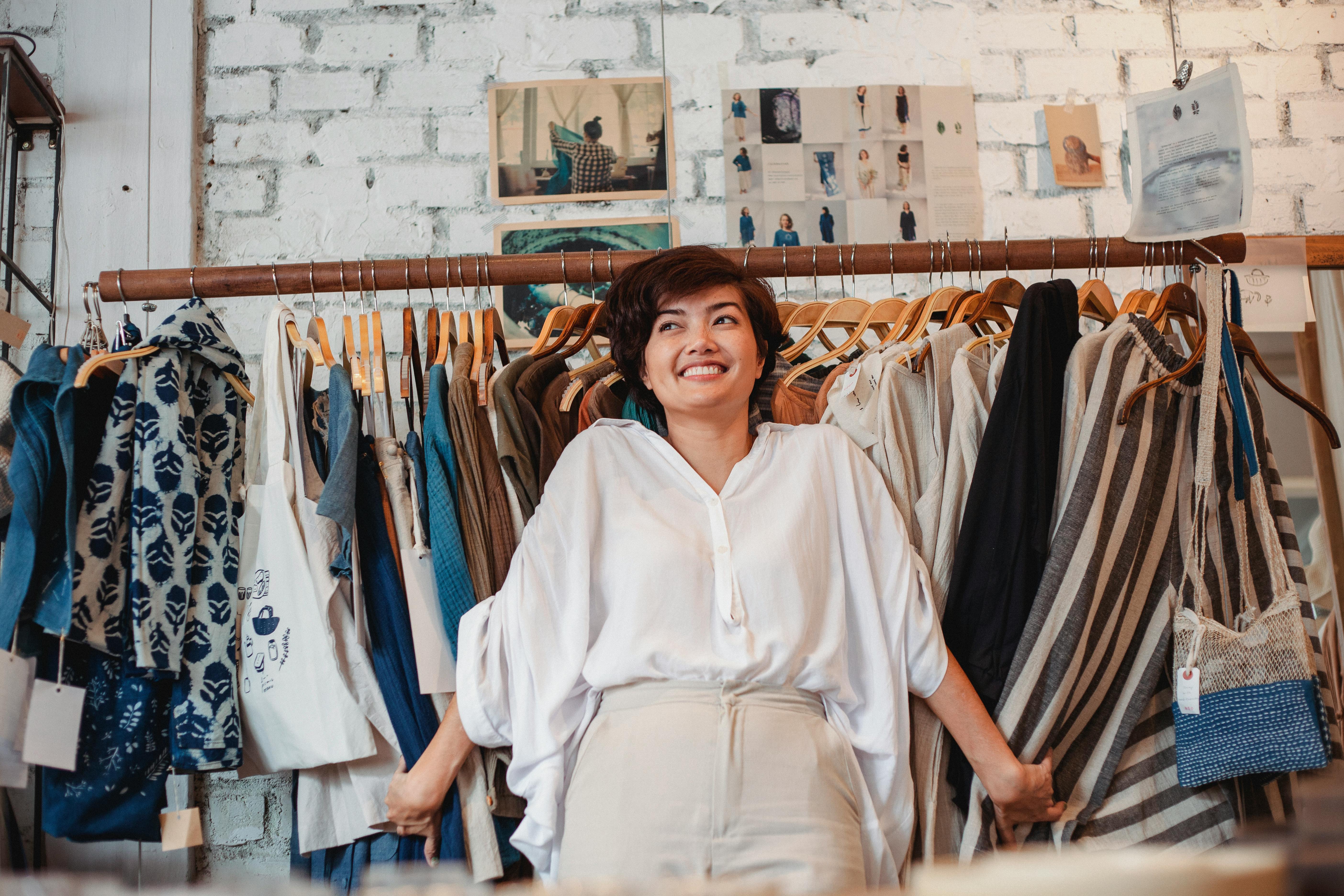 cheerful young ethnic lady smiling while holding stack of clothes hanging on rack
