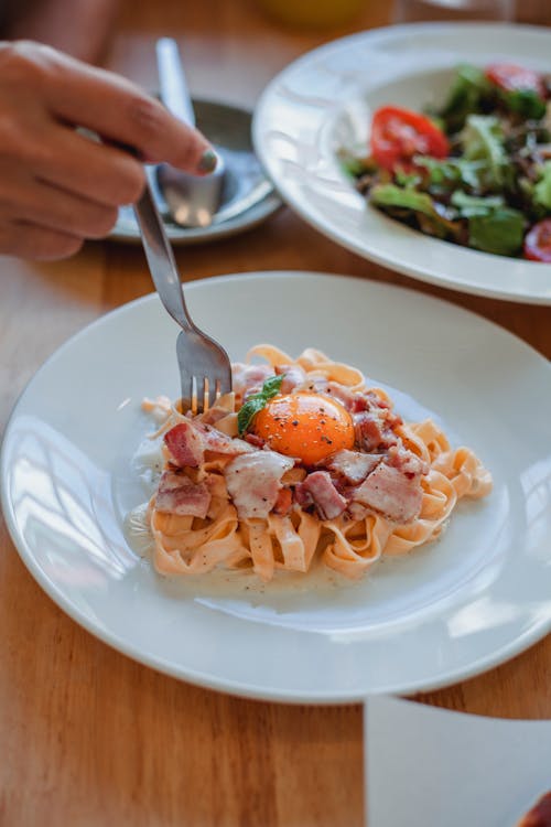 Free From above of unrecognizable person using fork to eat delicious pasta with bacon and egg in cafe during lunch time Stock Photo