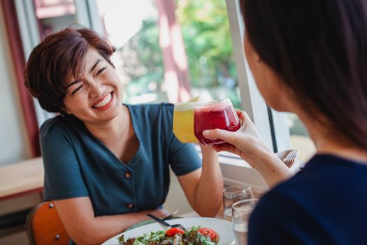 Cheerful Asian female with tilted head and short hair smiling and clinking glasses with crop friend while sitting in restaurant at table with salad