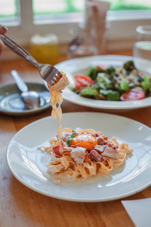 Fork and delicious pasta with bacon and spaghetti served on white plate near salad on table in restaurant with blurred background