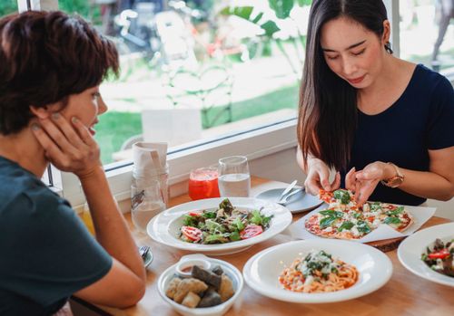 Free Content Asian woman taking slice of pizza while sitting at table near friend with delicious salad and pasta in restaurant Stock Photo