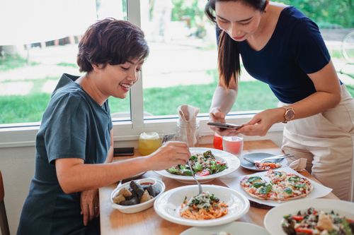 Free Happy Asian female taking picture on smartphone of delicious pasta while sitting in restaurant at served table with tasty dishes near friend Stock Photo