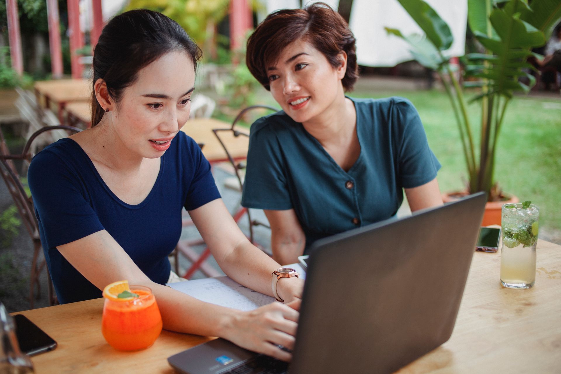 Content Asian female looking at busy coworker typing on computer while sitting at table and working online on veranda of cafe with blurred background