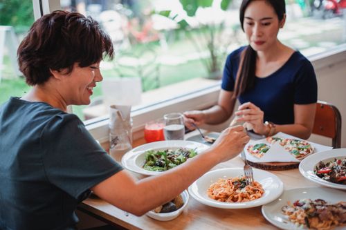 Free Asian female eating delicious pasta while sitting near female friend at served table with salad and pizza in restaurant during lunch Stock Photo