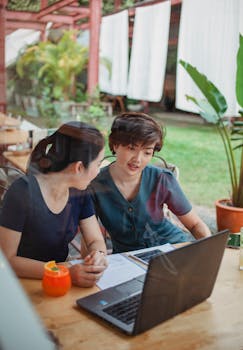 Two women collaborating at a café with laptops and documents in a casual outdoor setting.