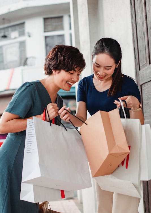 Ethnic female friends in casual clothes holding shopping bags and looking at goods while standing on street and smiling