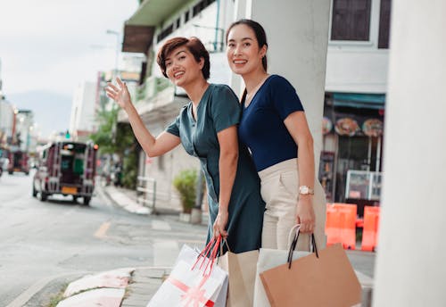 Low angle of smiling female friends with paper bags catching cab after shopping in mall