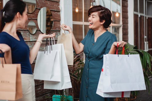 Laughing Asian female with shopping paper bags toothy smiling while standing with girlfriend near entrance of building