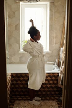 A woman in a bathrobe stretches by a bathtub in a sunlit bathroom.