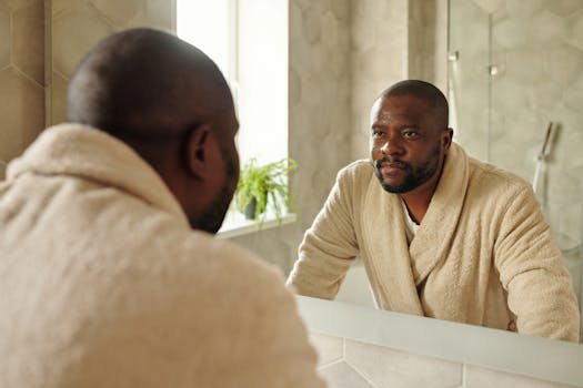 Adult man in a bathrobe looking at his reflection in a bathroom mirror. Morning routine concept.