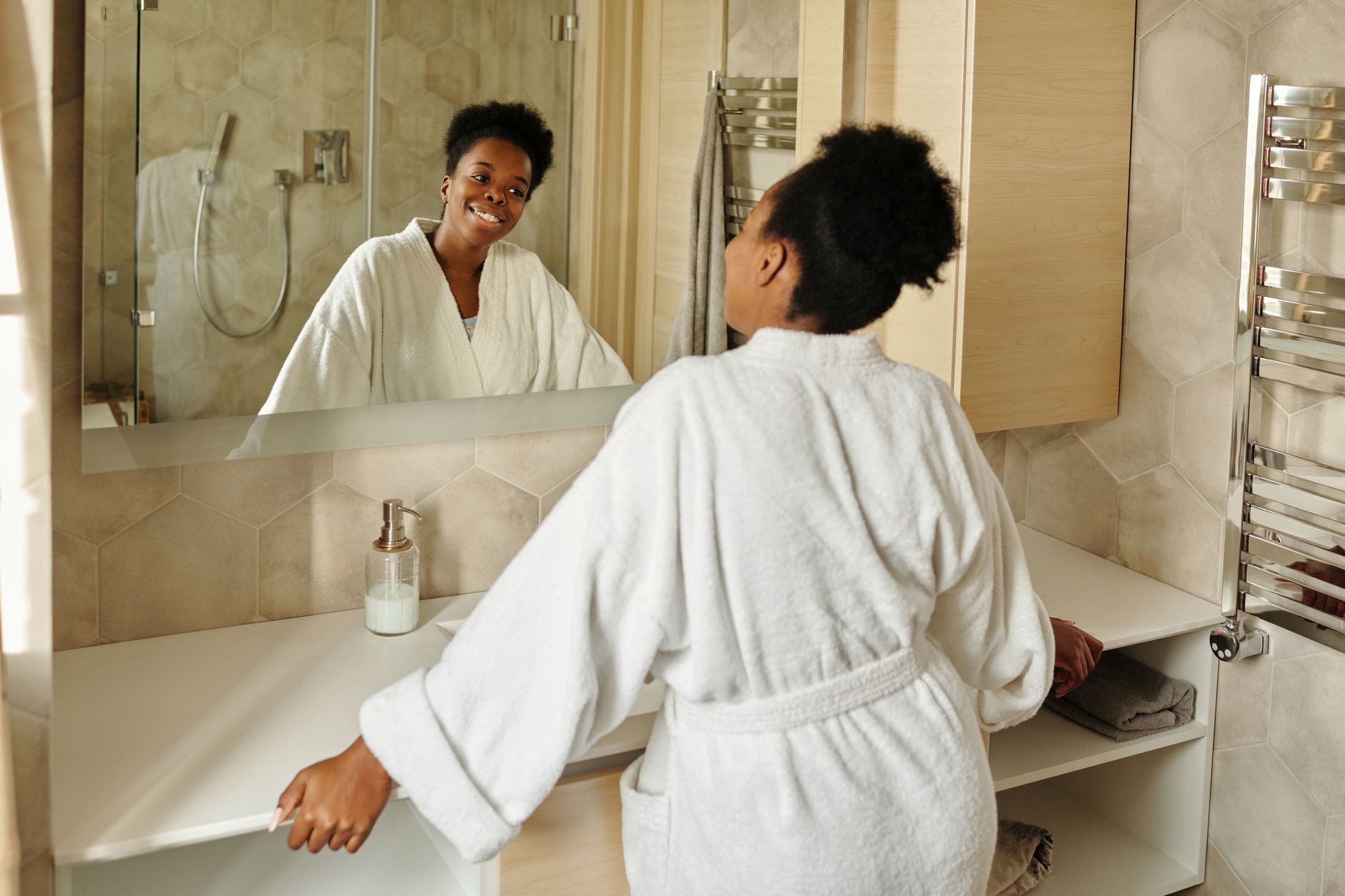 A woman in a bathrobe smiling at her reflection in a modern bathroom mirror.