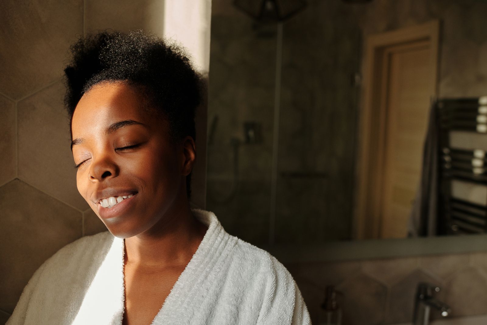 A serene woman in a bathrobe, eyes closed, enjoying sunlight indoors.
