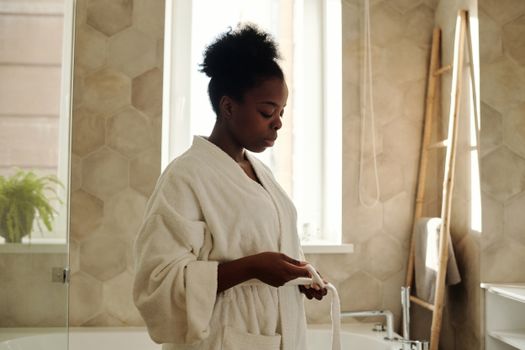 Woman wearing a bathrobe in a sunlit bathroom, enjoying a peaceful morning routine.