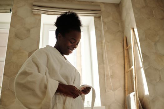 A joyful woman in a bathrobe smiles in a warmly lit bathroom, embracing relaxation.