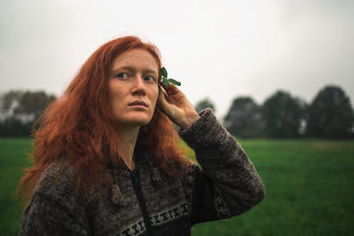 Contemplative female in casual wear decorating hair with green plant while standing on lush green meadow and looking away thoughtfully