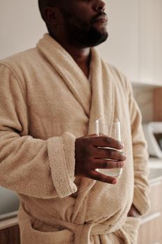 Adult man in a cozy bathrobe holding a glass of water while standing in a modern indoor setting.