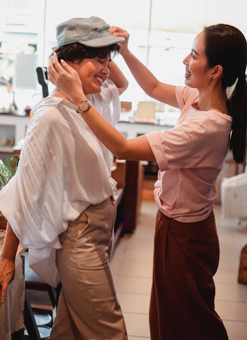 Side view of stylish female friends in trendy clothes standing in shop and trying on new hat