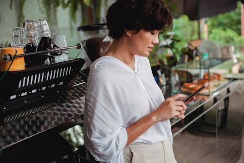 Free Side view of charming ethnic female in stylish clothes browsing mobile phone while leaning on professional coffee machine in cafe Stock Photo