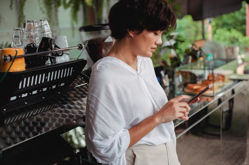 Side view of charming ethnic female in stylish clothes browsing mobile phone while leaning on professional coffee machine in cafe