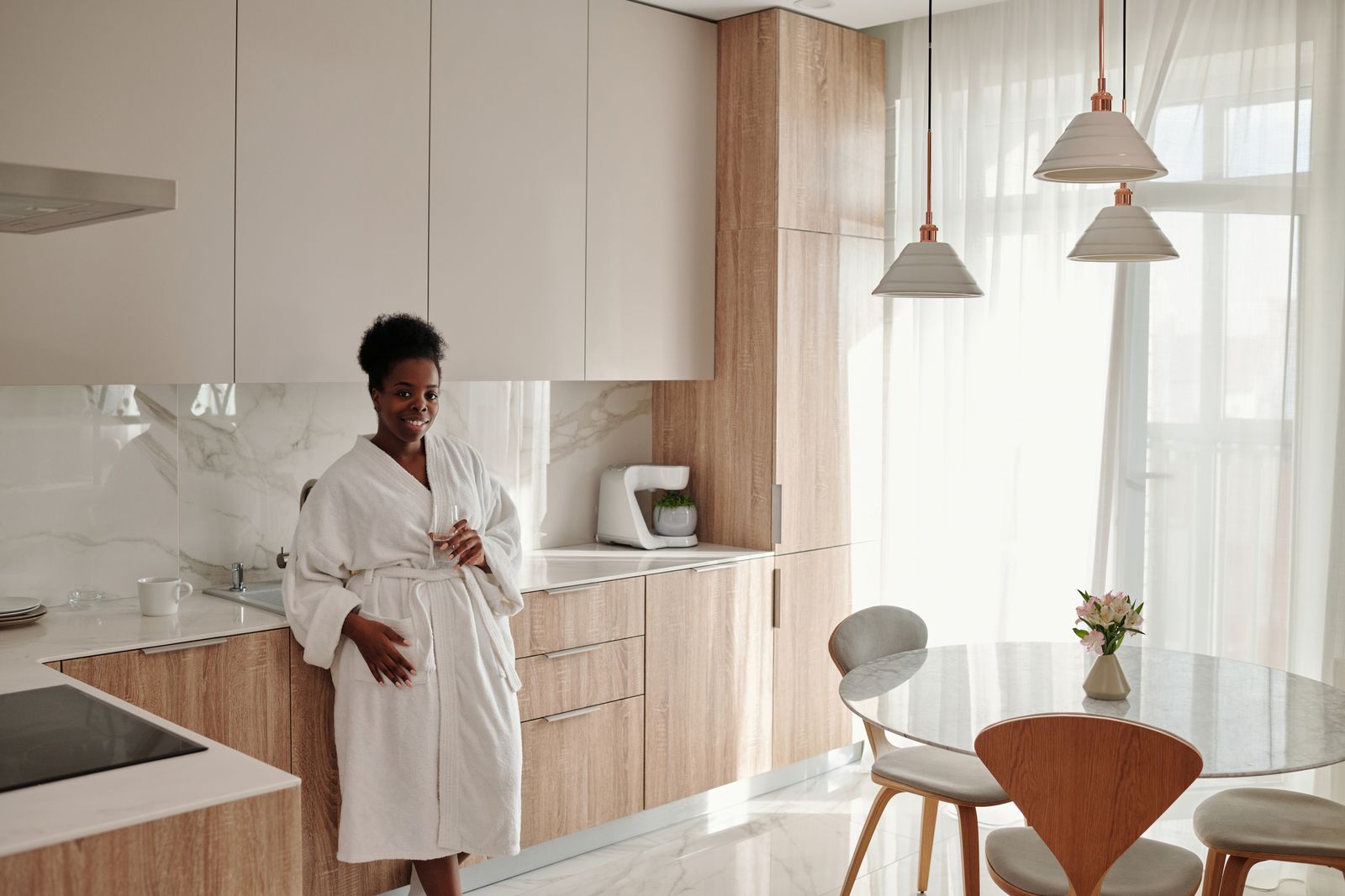 Smiling woman in a white bathrobe leaning against a counter in a sunlit modern kitchen