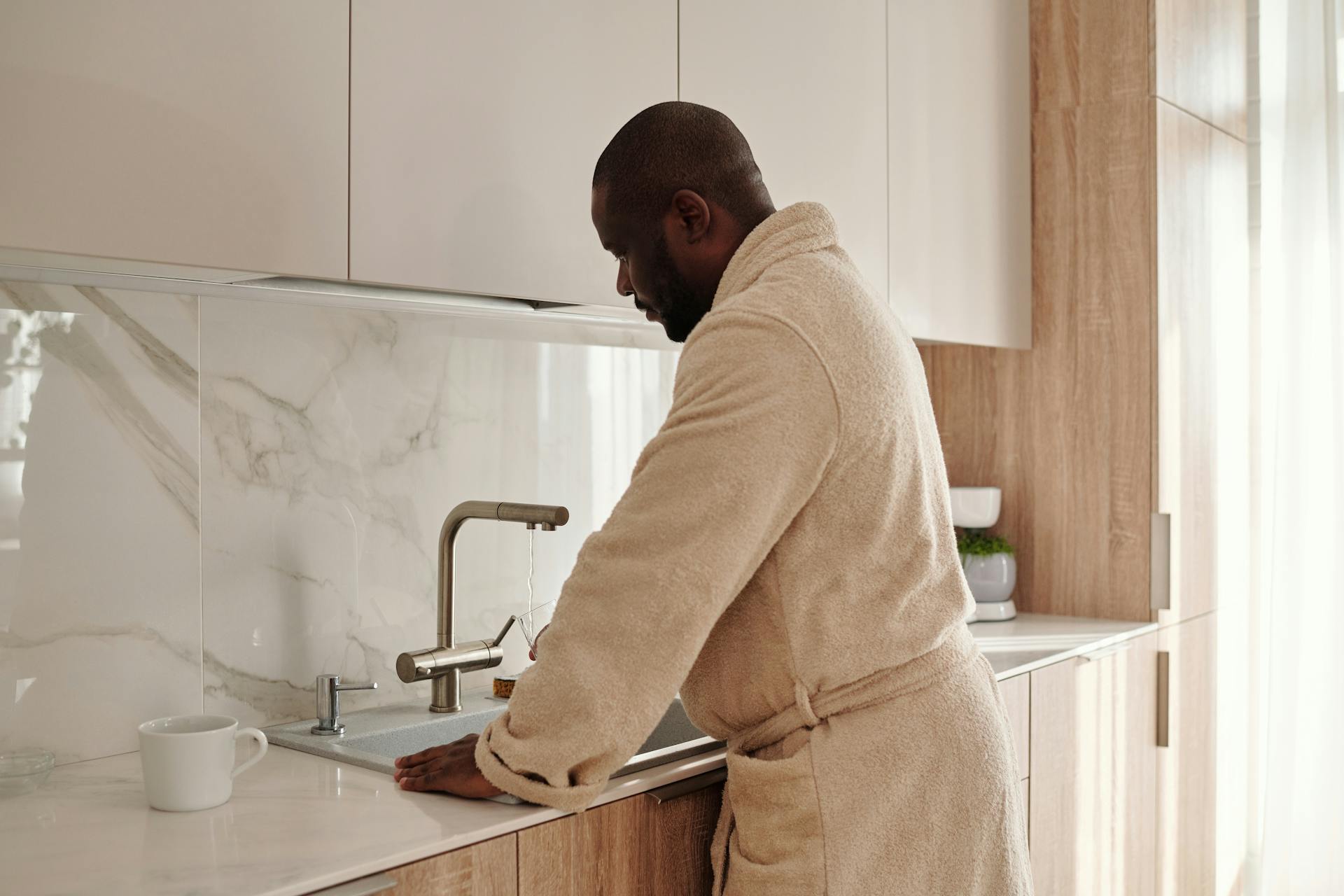 African American man in bathrobe washing hands at home sink in a modern kitchen.