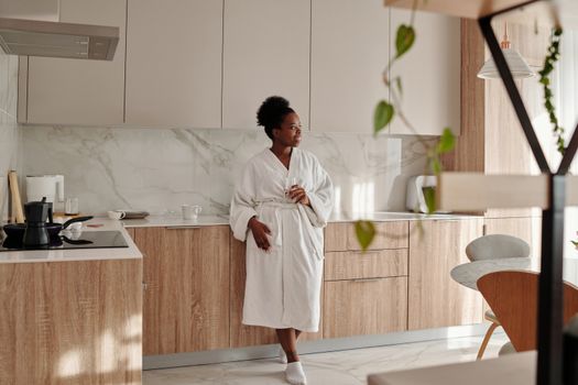 Black woman enjoying morning in a modern kitchen wearing a robe and drinking water.