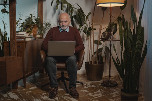 Elderly man working on a laptop in a cozy indoor home setting with plants and warm lighting.