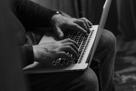 Monochrome image of a man in jeans typing on a laptop indoors.