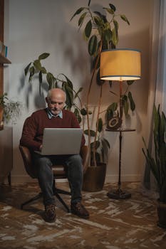 Elderly man sitting at home with laptop, surrounded by plants and cozy lighting.